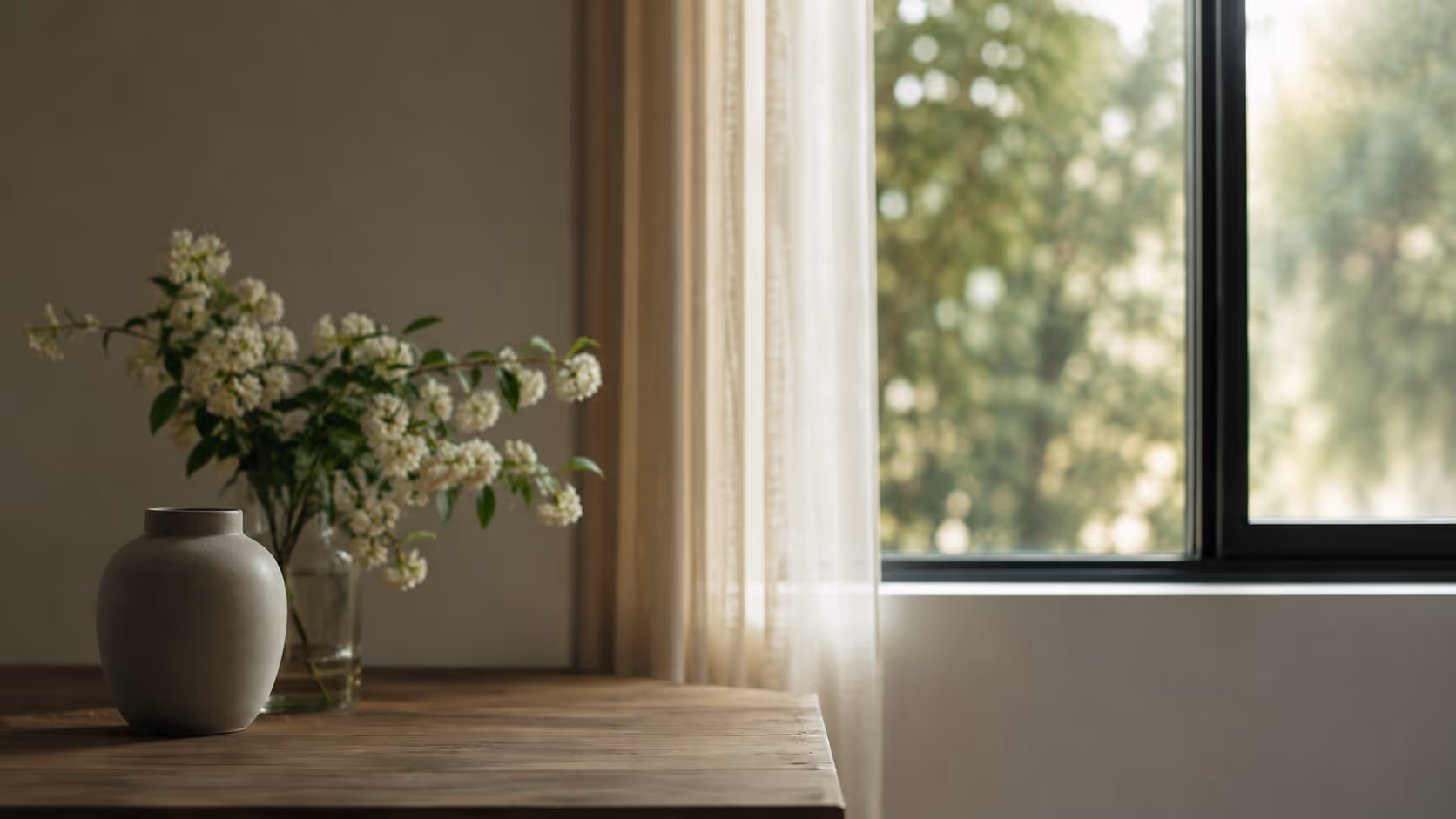 A pale ceramic urn beside a glass vase of white blossom on a wooden table by a sunlit window.